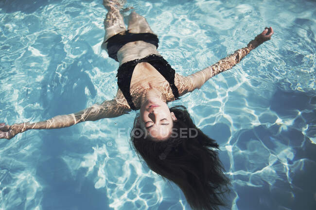 Serene woman in black bikini floating in sunny summer swimming pool — Stock Photo