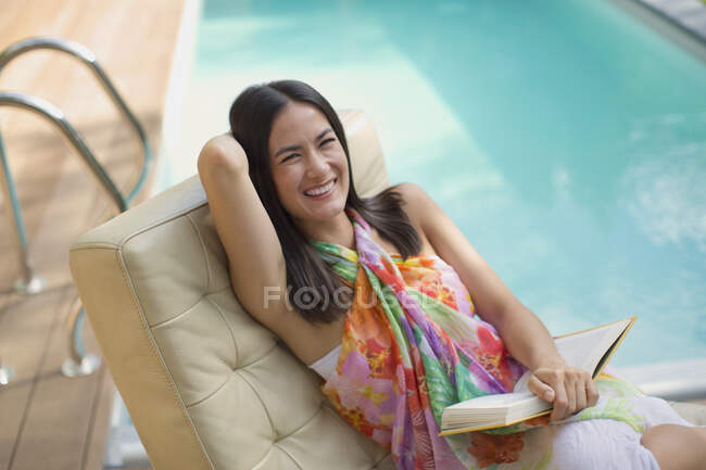 Portrait femme heureuse et riante lisant le livre sur chaise longue au bord de la piscine d'été — Photo de stock