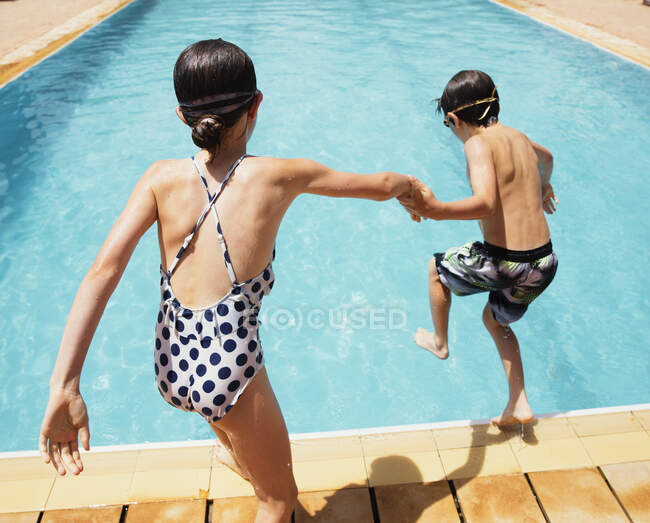 Niño y niña tomados de la mano, saltando a la soleada piscina de verano - foto de stock