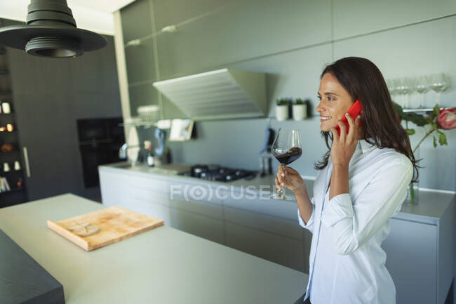 Mulher falando no telefone inteligente e beber vinho tinto na cozinha moderna — Fotografia de Stock