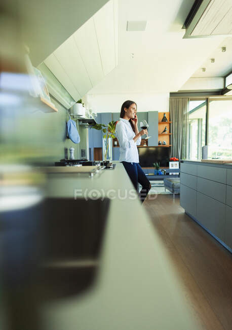 Жінка п'є червоне вино і розмовляє на смартфоні на сучасній кухні — стокове фото