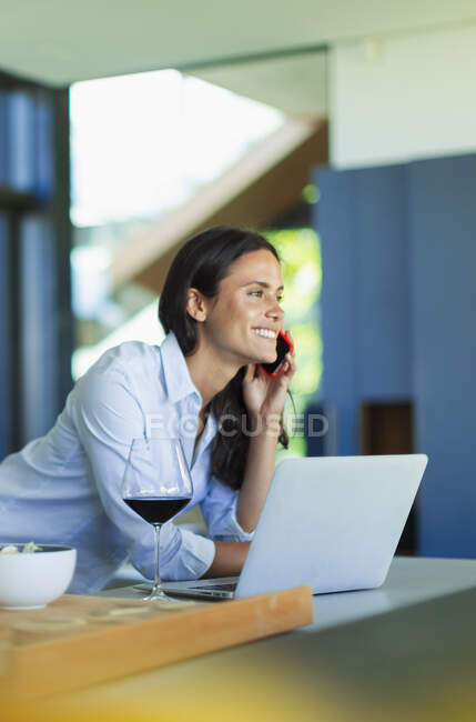 Усміхнена жінка розмовляє на смартфоні і п'є червоне вино на ноутбуці — стокове фото