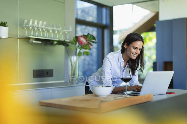 Усміхнена жінка використовує ноутбук і п'є червоне вино на кухні — стокове фото