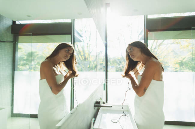 Woman wrapped in towel blow drying hair in sunny bathroom mirror — Stock Photo