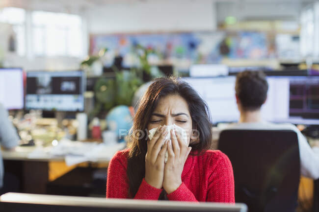 Mujer de negocios con alergias estornudando en el tejido en la computadora en la oficina - foto de stock