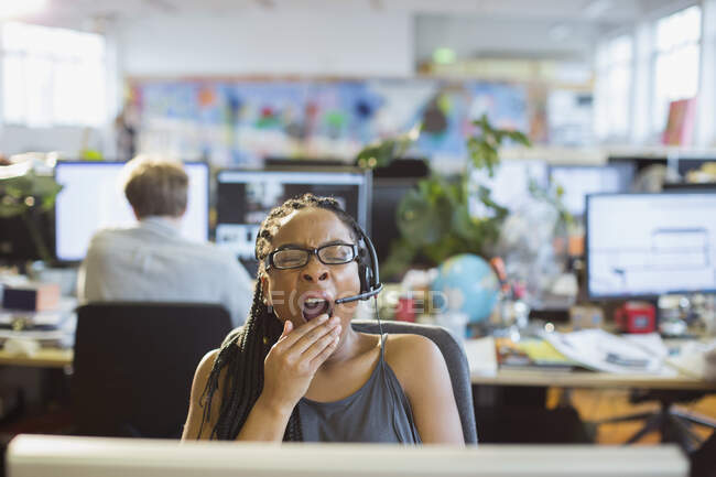 Уставшая деловая женщина с наушниками зевает на компьютер в офисе открытого плана — стоковое фото