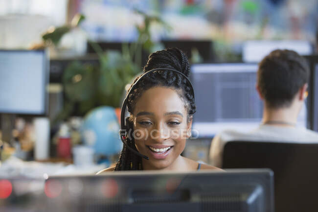 Улыбающаяся деловая женщина с наушниками, работающая за компьютером в офисе — стоковое фото