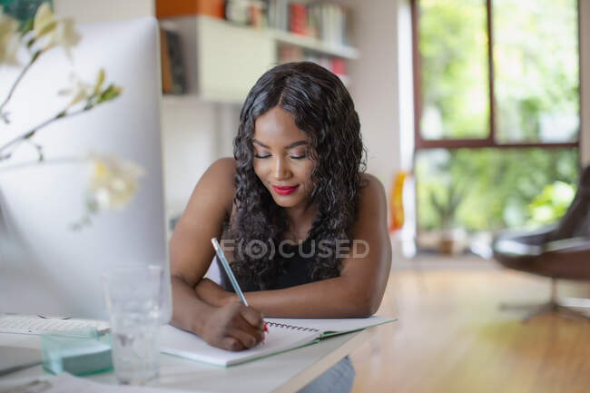 Mujer joven que escribe en cuaderno en el ordenador en la oficina de casa - foto de stock