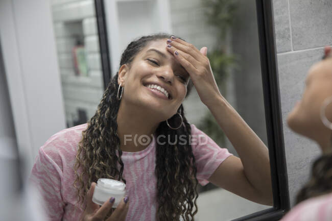 Happy young woman applying moisturizer in bathroom mirror. - foto de stock