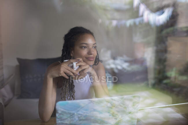 Pensar en la mujer joven bebiendo café en el ordenador portátil en la ventana - foto de stock