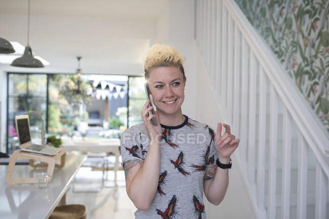 Усміхнена фрілансерка розмовляє на смартфоні на кухні — стокове фото