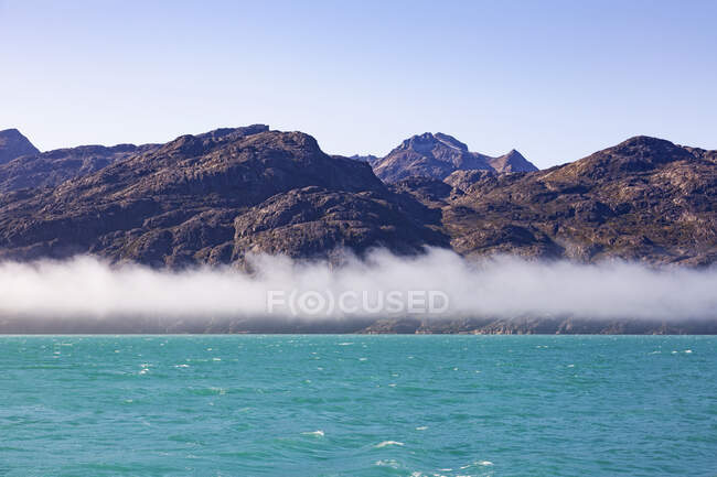 Nevoeiro abaixo de montanhas acidentadas e oceano azul-turquesa ensolarado Groenlândia — Fotografia de Stock