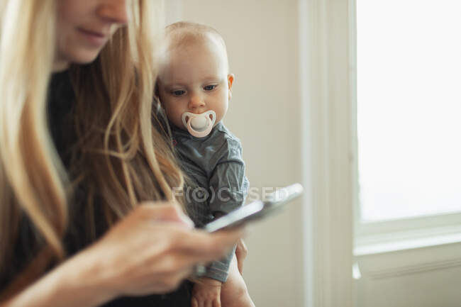 Hija curiosa con un pacificador viendo a la madre usando un teléfono inteligente. - foto de stock
