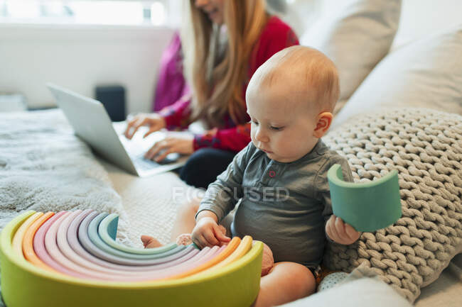 Curious baby girl playing with toy on bed while mother works at laptop — Stock Photo