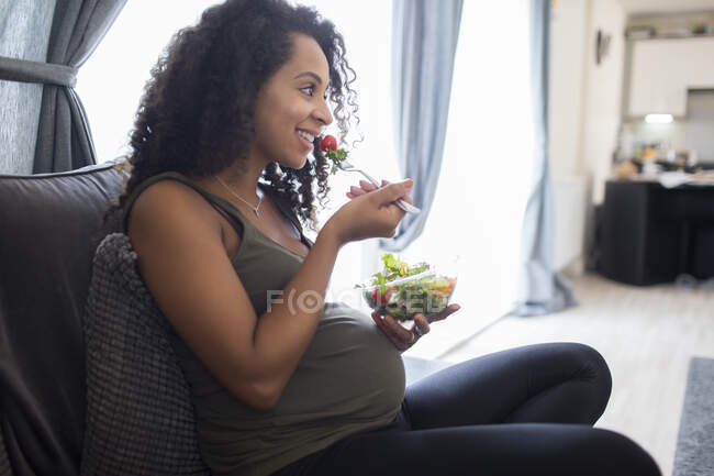 Sonriente joven embarazada comiendo ensalada en el sofá - foto de stock