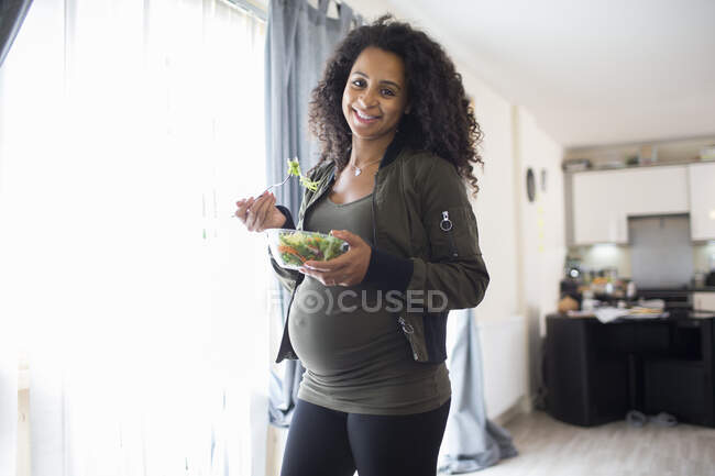 Retrato feliz joven embarazada comiendo ensalada en la ventana - foto de stock