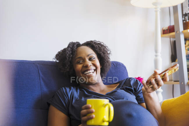 Lachende Schwangere mit Tee und Smartphone auf dem Sofa — Stockfoto