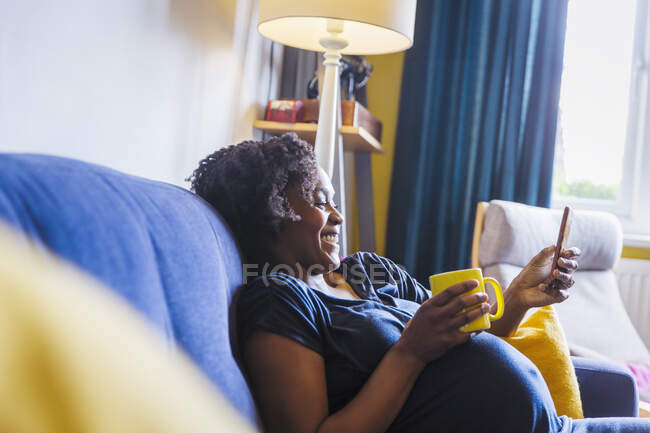 Счастливая беременная отдыхает на диване с чаем и смартфоном — стоковое фото