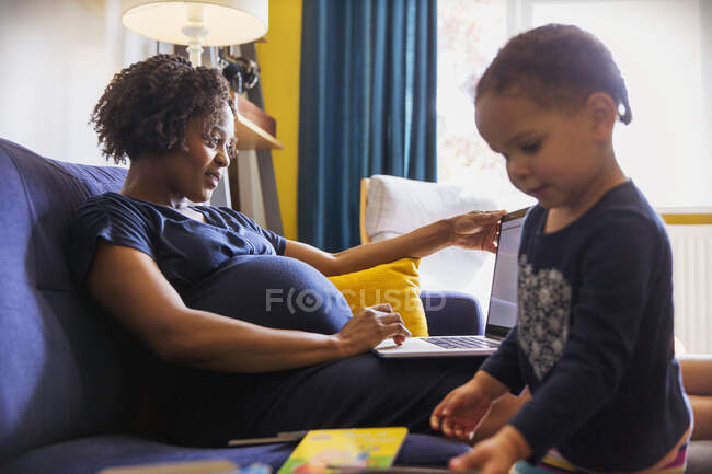 Schwangere Mutter benutzt Laptop, während Tochter auf Sofa spielt — Stockfoto