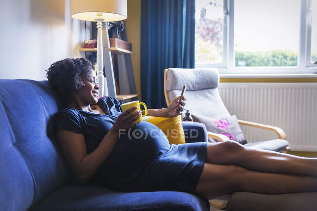 Mulher grávida feliz relaxando no sofá com chá e telefone inteligente — Fotografia de Stock