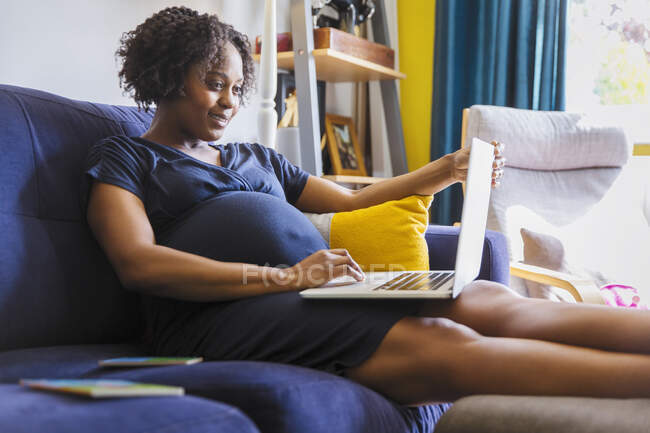 Femme enceinte utilisant un ordinateur portable sur un canapé — Photo de stock