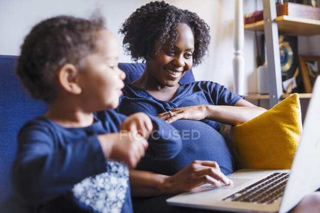 Счастливая беременная женщина с дочкой с ноутбуком на диване — стоковое фото