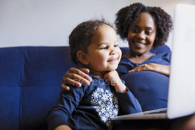 Mãe grávida feliz e filha pré-escolar usando laptop no sofá — Fotografia de Stock