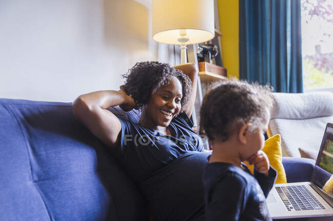 Счастливая беременная мать и дочь с ноутбуком на диване — стоковое фото