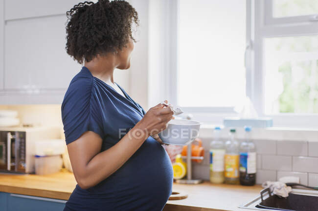 Pregnant woman eating and looking out kitchen window — Stock Photo