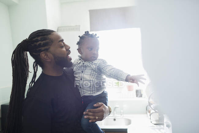 Pai com tranças longas segurando filho criança na cozinha ensolarada — Fotografia de Stock