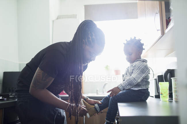 Père avec de longues tresses mettant des chaussures sur un tout-petit fils dans une cuisine ensoleillée — Photo de stock