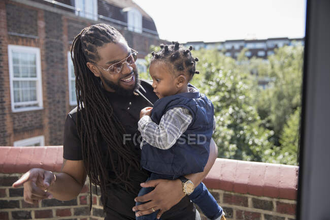 Батько з довгими плечима тримає малюка на сонячному балконі. — стокове фото