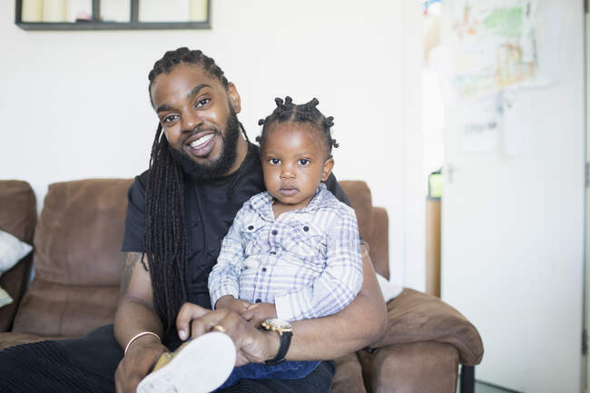 Portrait father with long braids putting shoes on toddler son on sofa — Stock Photo