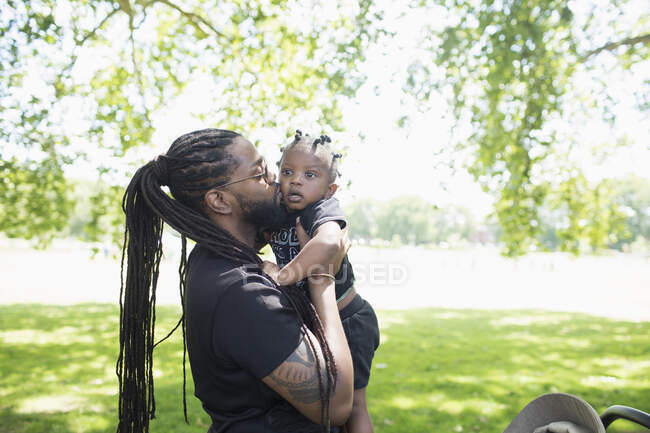 Батько з довгими косами цілує маленького сина в парку — стокове фото