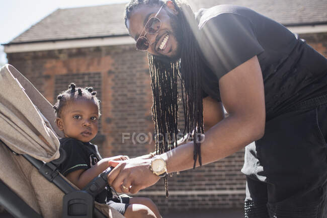 Portrait happy father with long braids fastening toddler son in stroller — Stock Photo