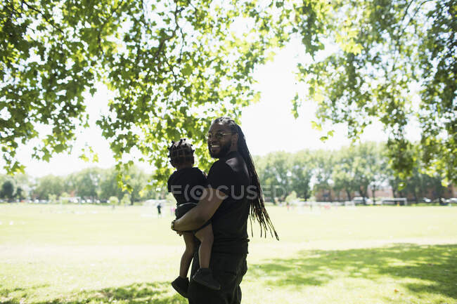 Retrato pai feliz com tranças longas carregando filho no parque ensolarado — Fotografia de Stock