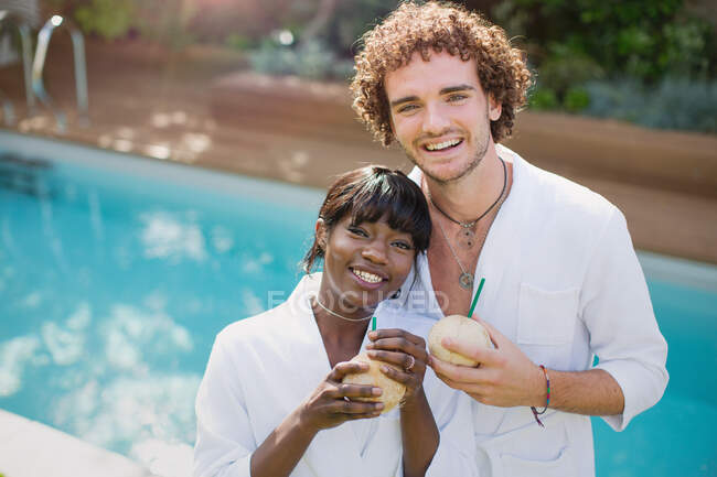 Portrait happy young couple in bathrobes drinking from coconuts at poolside — Stock Photo