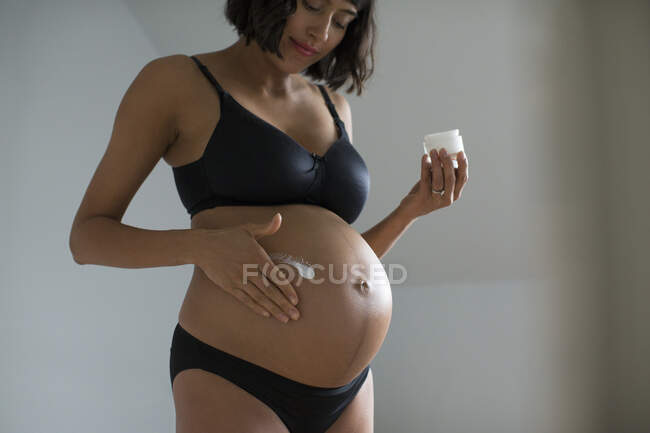 Беременная женщина в лифчике и трусиках наносит увлажняющий крем на живот — стоковое фото