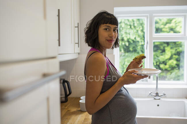 Retrato confiado mujer embarazada comiendo tostadas de aguacate en la cocina - foto de stock