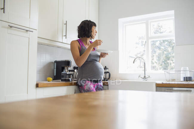Pensativa mujer embarazada comiendo en la cocina mirando por la ventana - foto de stock