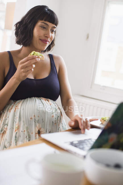 Беременная женщина ест за ноутбуком — стоковое фото