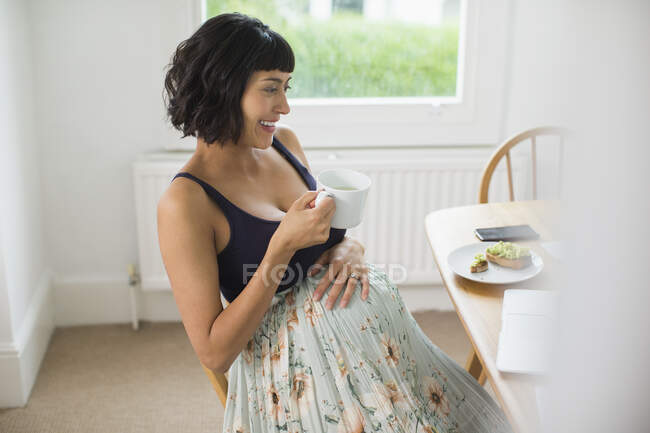 Счастливая беременная женщина пьет чай за ноутбуком — стоковое фото