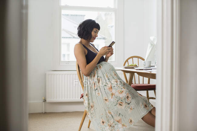Mulher grávida em vestido floral usando telefone inteligente na mesa — Fotografia de Stock