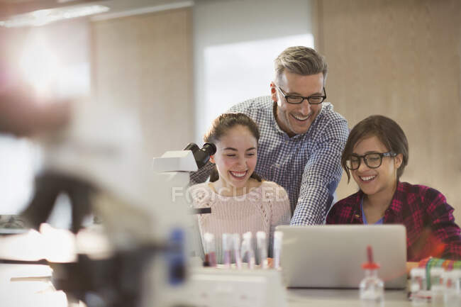 Profesora y alumna masculina realizando experimentos científicos al microscopio y portátil en aula de laboratorio - foto de stock