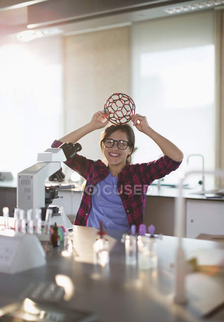 Портрет грайливої студентки, що тримає молекулярну структуру на голові в мікроскопі в лабораторному класі — стокове фото