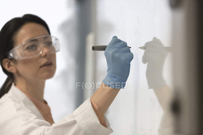 Female science teacher wearing lab coat, rubber glove and goggles, writing at whiteboard in classroom — Stock Photo