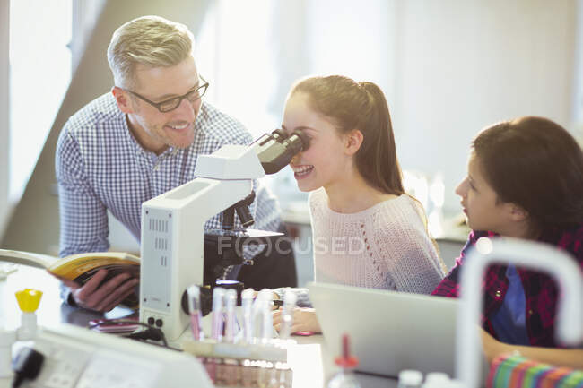 Учитель-мужчина помогает ученице с помощью микроскопа, проводит научный эксперимент в лабораторном классе — стоковое фото