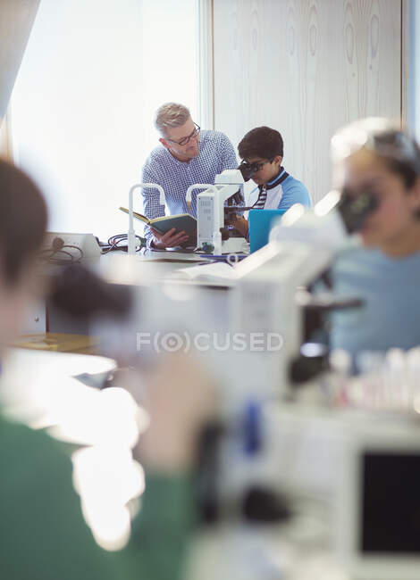 Profesor y estudiante masculino usando microscopio, llevando a cabo experimento científico en aula de laboratorio - foto de stock