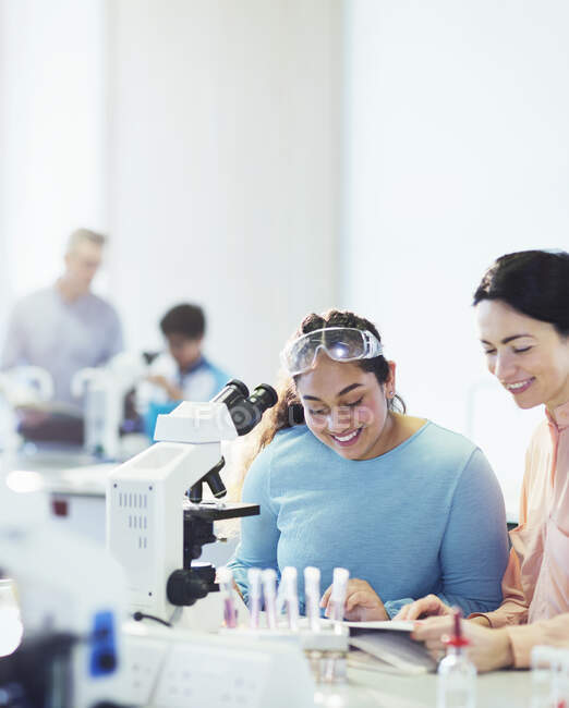 Professora de ciências do sexo feminino sorridente e estudante lendo o livro didático ao microscópio em sala de aula de laboratório — Fotografia de Stock