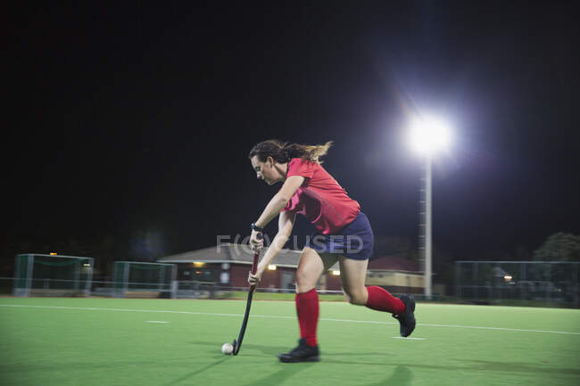 Entschlossene junge Hockeyspielerin läuft mit Hockeyschläger und Ball, spielt nachts auf dem Feld — Stockfoto